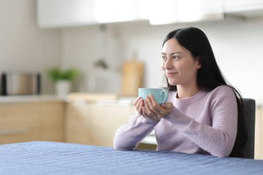 Rahatlamış Asyalı kadın evdeki mutfakta içiyor ve düşünüyor.