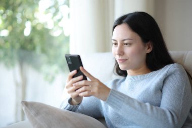 Evdeki koltukta oturan Asyalı kadın akıllı telefon kullanıyor.