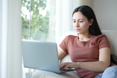 Ciddi Asyalı kadın evdeki koltukta dizüstü bilgisayar kullanıyor.