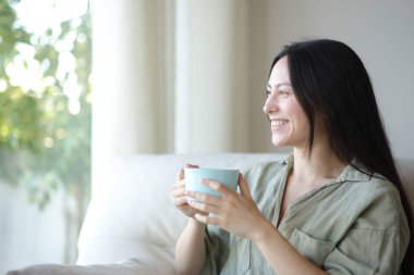 Mutlu Asyalı kadın kahve içiyor ve pencereden bakıyor. Evdeki kanepede oturuyor.