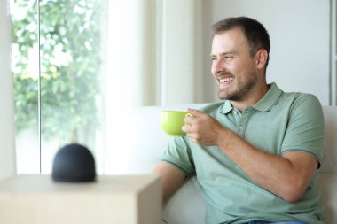 Mutlu bir adam kahve içiyor akıllı bir hoparlörle evde bir kanepede oturuyor.