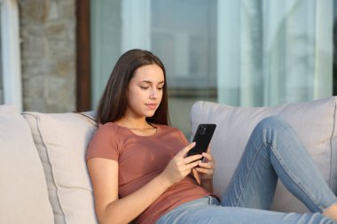 Balkonda rahat bir kadın cep telefonunu kontrol ediyor evdeki kanepede oturuyor.