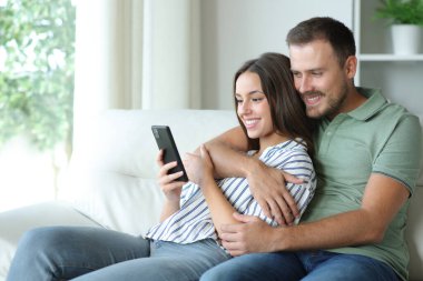 Mutlu çift cep telefonu kullanıyor. Evdeki koltukta oturuyor.