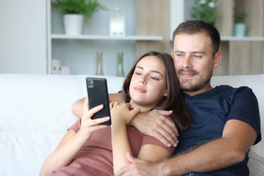 Ciddi bir çift telefonlarını kontrol edip evdeki koltukta oturuyor.