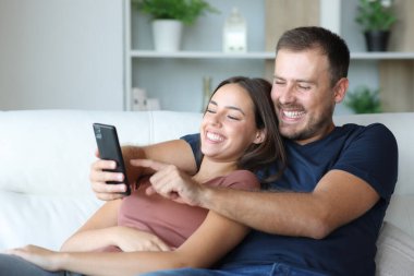 Mutlu çift cep telefonu kullanıyor. Evdeki koltukta oturuyorlar.