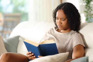 Evdeki kanepede oturup kötü bir kağıt kitap okuyan hayal kırıklığına uğramış zenci kadın.