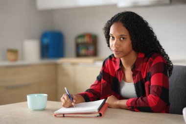 Ciddi siyahi öğrenci evdeki bir mutfakta sana bakarak notlar yazıyor.