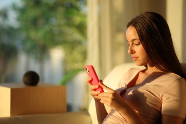 Ciddi bir kadın gün batımında akıllı hoparlörün yanında telefon kullanıyor. Evdeki kanepede oturuyor.