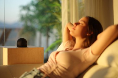 Gün batımında akıllı hoparlörlerin yanında rahat bir kadın evdeki kanepede oturuyor.