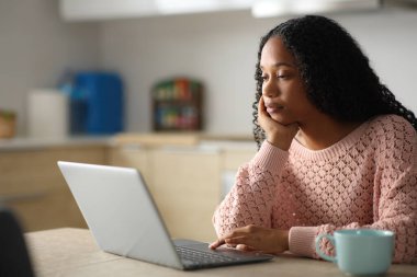 Ciddi siyahi kadın evdeki bir mutfakta kişisel bilgisayar kullanıyor.