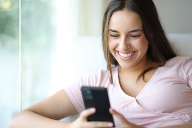 Evin içindeki mutlu kadın koltukta oturan cep telefonunu kullanıyor. 