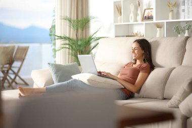 Evdeki oturma odasında dizüstü bilgisayar kullanan mutlu bir kadın.