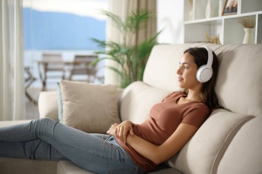 Ciddi bir kadın, kulaklık takıyor, müzik dinliyor evdeki bir salonda, bir kanepede oturuyor.
