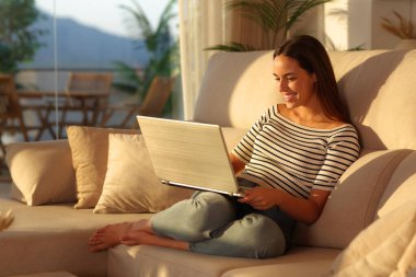 Gün batımında evinde dizüstü bilgisayarla koltukta oturan mutlu kadın.