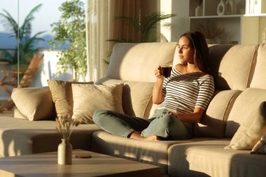 Kanepede oturup kahve içen rahat bir kadın evdeki pencereden dışarı bakıyor.