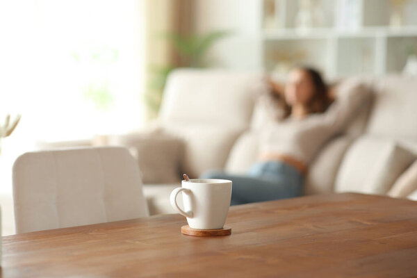 Coffee cup in foreground on a wooden table and woman relaxing sitting on a couch in the background at home