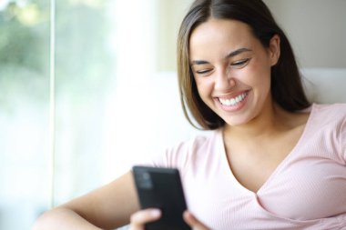 Mutlu kadın cep telefonuyla gülerken evdeki koltukta oturuyor.