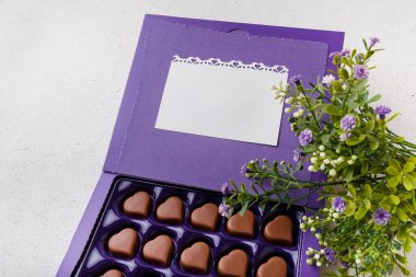 Mor kalp şeklinde çikolatalar, kartpostallar ve beyaz arka planda çiçekler. Sevgililer Günü 14 Şubat tatlı hediye