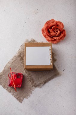 Hediye kutusu, kumaş gül çiçeği, boş kartpostal, çuval bezi ve beyaz arka planda kalp şeklinde kırmızı mum. Sevgililer günü, doğum günü, düğün