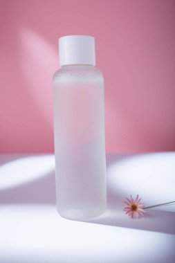 Mock up bottle of essence toner with no label in trendy natural light with flower on pink backdrop. Face skin care cosmetics, moisturizer, copy space