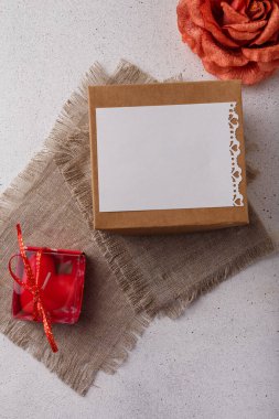 Hediye kutusu, kumaş gül çiçeği, boş kartpostal, çuval bezi ve beyaz arka planda kalp şeklinde kırmızı mum. Sevgililer günü, doğum günü, düğün