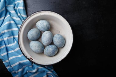 Easter eggs painted blue in white bowl on black wooden background with striped fabric. Top view, copy space