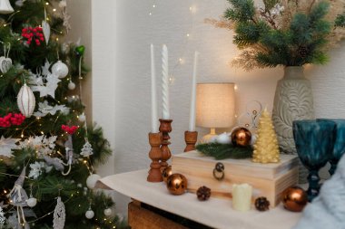Kahverengi ampullü ahşap kutu, köknar dalı mumlar ve köknar ağacı şeklinde altın mum. Arka planda Noel çelengi ışıkları.