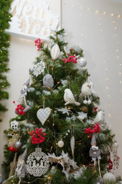 Beyaz kuşlar, tahta oyuncaklar ve kırmızı böğürtlenlerle süslenmiş güzel Noel ağacı. Arka planda çelenk ışıkları var. Dikey yakın çekim.