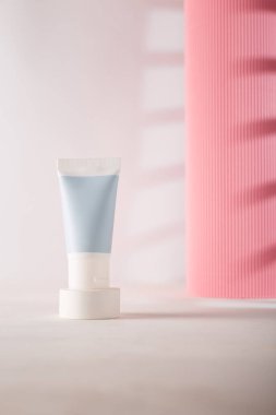 Beyaz beton kaidede yumuşak mavi kozmetik tüp, arka planda pembe kağıt rulo. Boş etiket, damgalanmamış şablon, model. Uzayı kopyala Ürün reklam afişi.