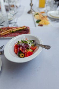 Beyaz tabakta Yunan salatası, bezelye yeşiliyle süslenmiş aperatif. Beyaz masa arkasında biraz et ve peynir tabağı. Şölen aperatifleri