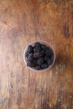 Olgun böğürtlenler kahverengi eski ahşap masa arkasında plastik bardakta, yakın çekim. Üst görünüm, alanı kopyala.