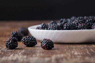 Beyaz tabağın bir kısmı olgun taze böğürtlenlerle ahşap eski kırsal masa arka planında görülebiliyor. Yakınlarda böğürtlen. Yakın çekim, boşluğu kopyala.