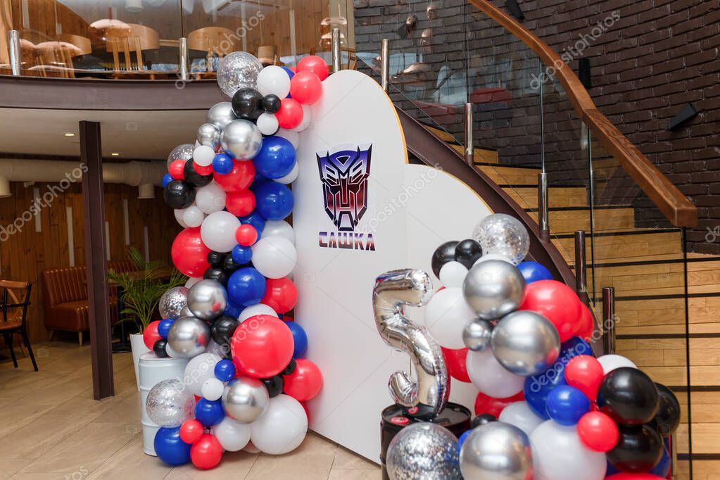 Izmail, Ukraine. 21st July, 2024. Multicolor balloons decoration for 5 years old boy birthday party. Photo zone with Transformers logo on white background. Sideview. Inscription - Sashka (name)