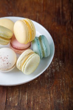 Beyaz tabakta farklı tatlara sahip çok renkli badem kurabiyeleri, eski ahşap masa arkaplanı, yakın çekim üst görüntü.