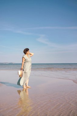 Yaz günbatımında tropikal ada sahilinde yürüyen uzun elbiseli mutlu yaşlı kadın. Emekli yaşlı kadın dinleniyor ve yaz tatilinde açık hava aktivitelerinin tadını çıkarıyor..