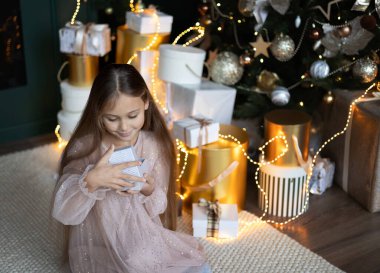 Uzun elbiseli şirin küçük kız yerde oturuyor ve arka plandaki Noel ağacı için bir hediye kutusu açıyor. Xmas tatil konsepti