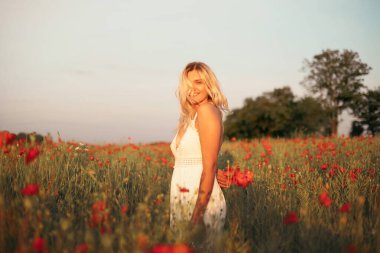 Gün batımında haşhaş tarlasında beyaz elbiseli güzel sarışın kadın. Kadın ve doğa