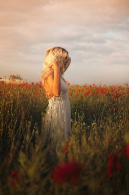 Gün batımında haşhaş tarlasında beyaz elbiseli güzel sarışın kadın. Kadın ve doğa