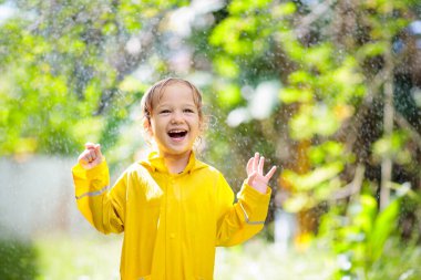 Güneşli sonbahar gününde yağmurda oynayan bir çocuk. Çocuk ağır duş altında ve sarı ördek şemsiyesiyle. Ördek su geçirmez ayakkabılı küçük çocuk. Lastik çizmeler. Yağmurlu havaya göre açık hava aktivitesi