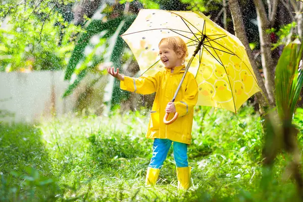 Child playing in the rain on sunny autumn day. Kid under heavy shower with yellow duck umbrella. Little boy with duckling waterproof shoes. Rubber wellies boots. Fall outdoor activity by rainy weather