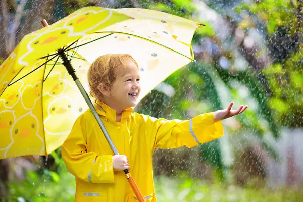 子供は晴れた秋の日に雨の中で遊ぶ 黄色いアヒルの傘で重いシャワーの下の子供 小さな男の子とアヒルの防水靴 ラバーウェリーブーツ 雨天による屋外活動 ストックフォト