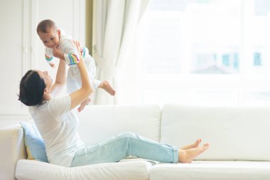 Anne ve çocuk beyaz bir kanepede. Asyalı anne ve oğlan güneşli yatak odasında oynuyorlar. Ebeveyn ve küçük çocuk evde dinleniyor. Aile birlikte eğleniyor. Bebek odası için yatak ve tekstil