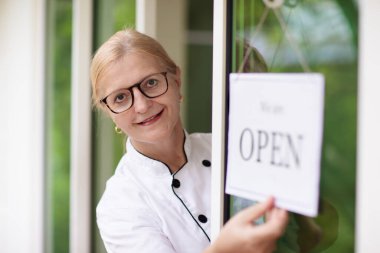 Kafe açılış töreni. Restoranın ya da marketin ön kapısında açık tabela var. Önlüklü garson müşteriyi selamlıyor. Pastane girişinde hoş geldin mesajı. 