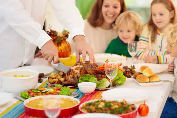 感謝祭の夕食を食べる子供たちと家族 秋の装飾が施されたダイニングテーブルの上で七面鳥とカボチャのパイをロースト お祝いの食事をしている親と子供 お父さんとお母さんが肉を切る ストック写真