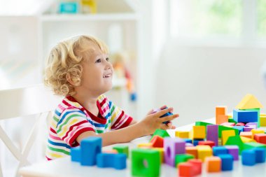 Çocuk renkli oyuncak bloklarıyla oynuyor. Küçük çocuk oyuncaklardan kule inşa ediyor. Küçük çocuklar için eğitici ve yaratıcı oyuncaklar ve oyunlar. Gökkuşağı tuğlalı beyaz yatak odasındaki bebek. Evdeki çocuk.