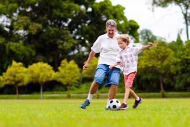 Baba ve oğlu dışarıda futbol oynuyor. Baba ve çocuk park stadyumunda koşuyor. Adam ve küçük çocuk güneşli yaz gününde futbol oynuyorlar. Genç aile için sağlıklı aktif spor.