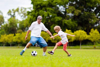 Baba ve oğlu dışarıda futbol oynuyor. Baba ve çocuk park stadyumunda koşuyor. Adam ve küçük çocuk güneşli yaz gününde futbol oynuyorlar. Genç aile için sağlıklı aktif spor.