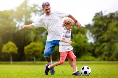 Baba ve oğlu dışarıda futbol oynuyor. Baba ve çocuk park stadyumunda koşuyor. Adam ve küçük çocuk güneşli yaz gününde futbol oynuyorlar. Genç aile için sağlıklı aktif spor.