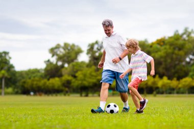 Baba ve oğlu dışarıda futbol oynuyor. Baba ve çocuk park stadyumunda koşuyor. Adam ve küçük çocuk güneşli yaz gününde futbol oynuyorlar. Genç aile için sağlıklı aktif spor.