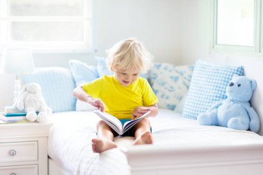 Beyaz güneşli bir odada pencereli bir çocuk yatakta oynuyor. Çocuk odası ve iç tasarım. Erkek bebek evde. Çocuk yuvası için yatak ve tekstil. Oyuncağı ve kitabı olan bir çocuk. Uyku ve uyku zamanı..